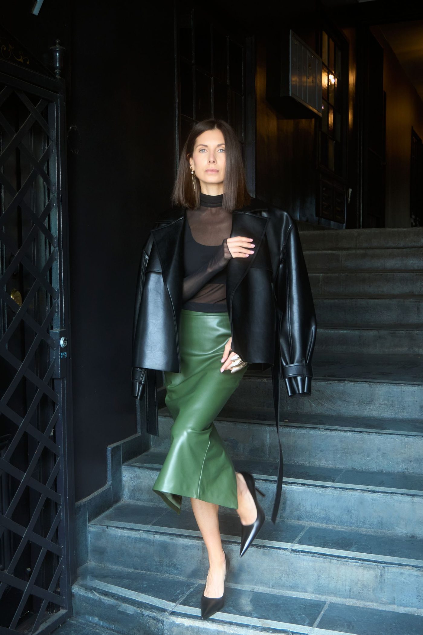 Valeriya on a dark stone stairway in a black leather jacket, sheer black top and olive leather skirt